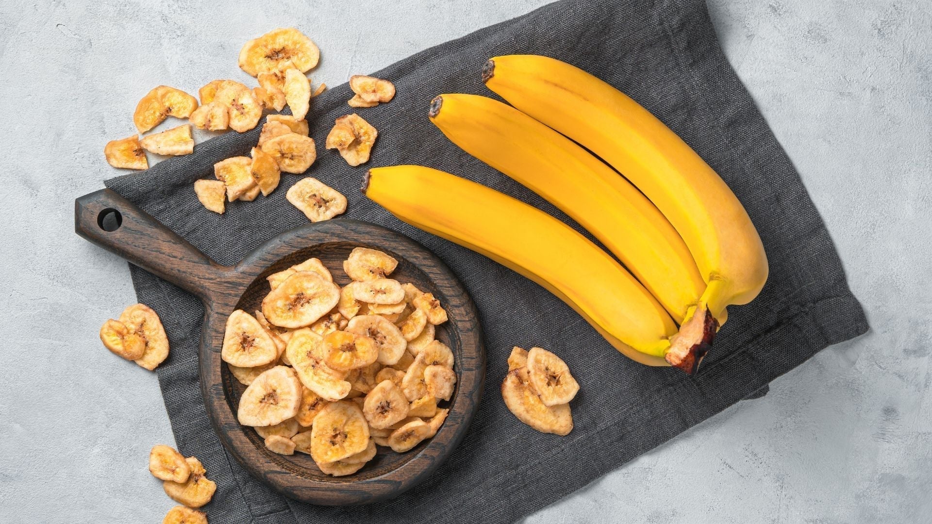 Bowl of dried banana chips on wooden table with cloth, showcasing yellow, ripe, organic tropical fruit snacks rich in potassium.